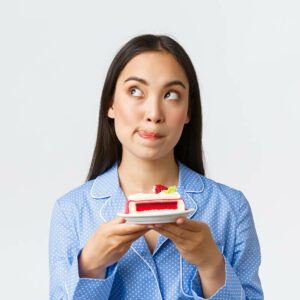 close up shot thoughtful cute asian girl standing night pajama with plate cake looking upper left corner deciding cant resist temptation eat delicious dessert white background